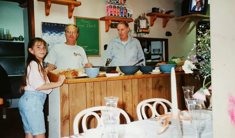 Two men and a young girl behind the wooden serving counter at Rose's Sugar Shack during a Thanksgiving spread, the cafe's chalkboard sign on the wall behind them