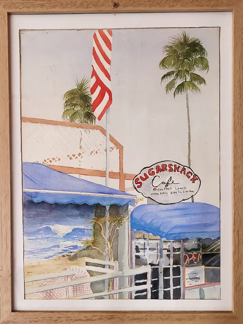 Framed watercolor painting of the Sugar Shack Cafe storefront with its oval sign, blue awnings, American flag, palm trees and an ocean wave in the background