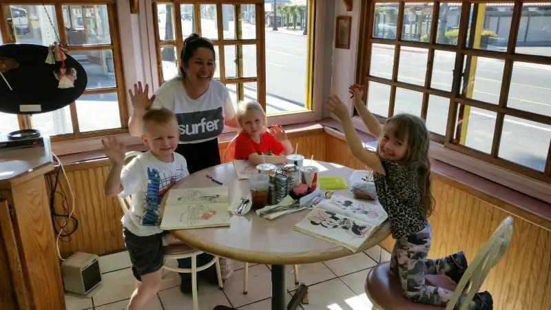 A mother and three young children waving and smiling over coloring pages at a sunny corner window table