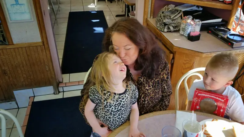 A woman kissing a young girl on the forehead at the table while a young boy looks on