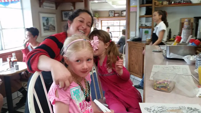 A woman hugging two young girls in pink at a table while a server works the counter behind them in a busy cafe