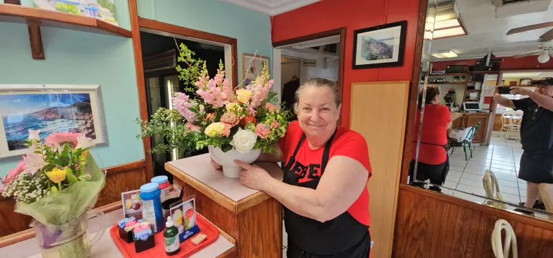 Owner smiling behind the counter of Rose's Sugar Shack Cafe with a floral bouquet, 2024