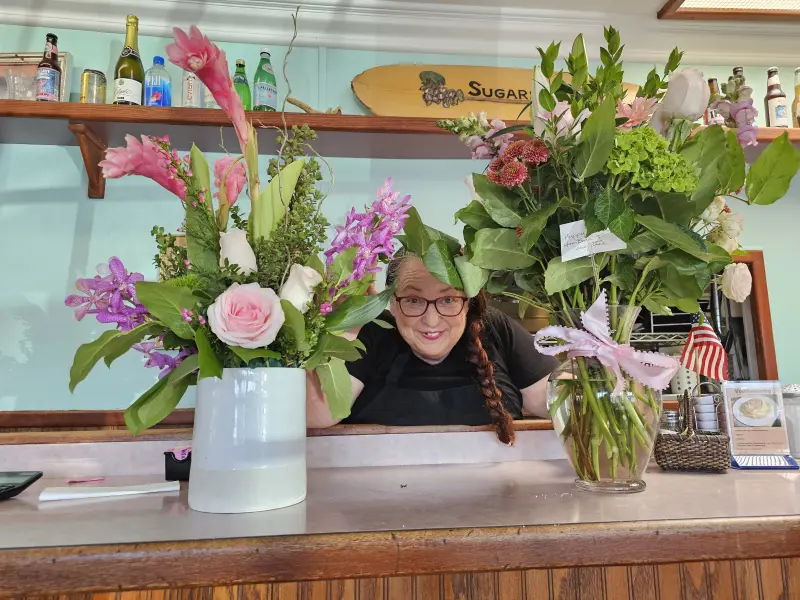 Owner smiling between two large flower bouquets at the counter of Rose's Sugar Shack Cafe, 2025