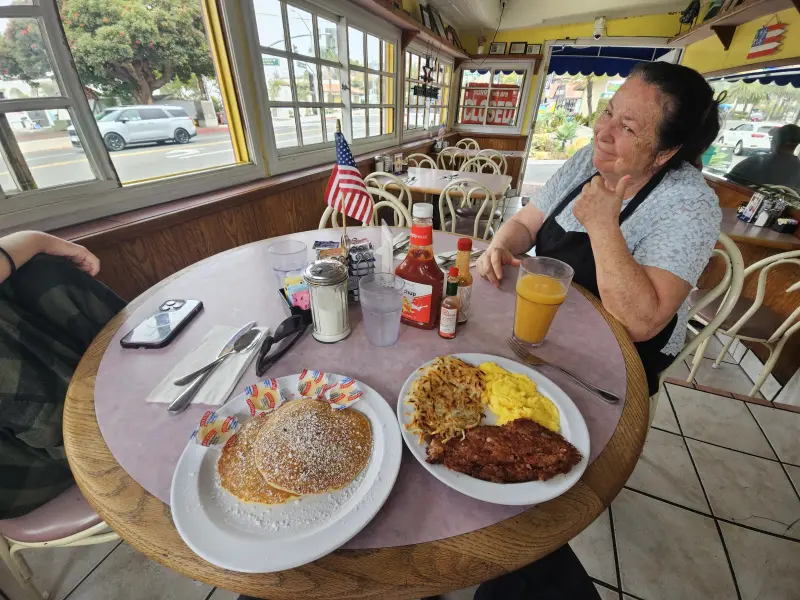 Owner giving a thumbs-up beside a breakfast of pancakes, eggs, hash browns and country-fried steak at Rose's Sugar Shack Cafe, 2025