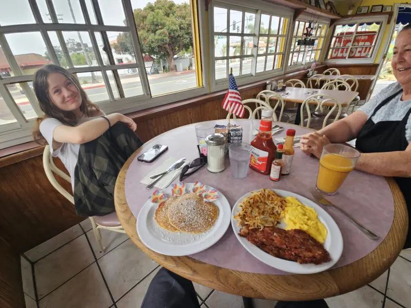 A young girl and the owner at a window table with a big breakfast spread at Rose's Sugar Shack Cafe, 2025