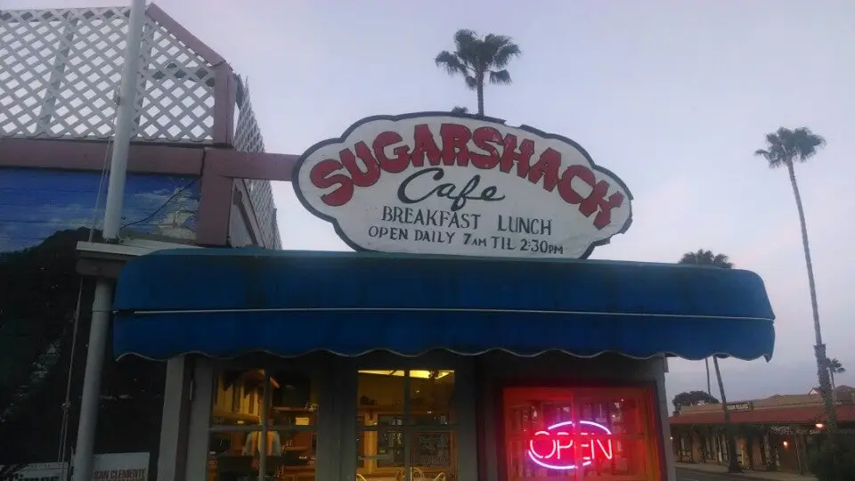 Hand-painted Sugarshack Cafe Breakfast Lunch sign at dusk with a glowing neon Open sign and palm trees