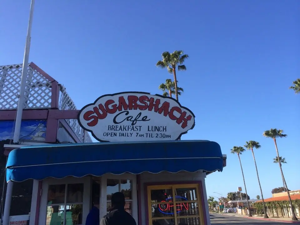 Sugarshack Cafe hand-painted sign against a clear blue sky with palm trees and a blue awning