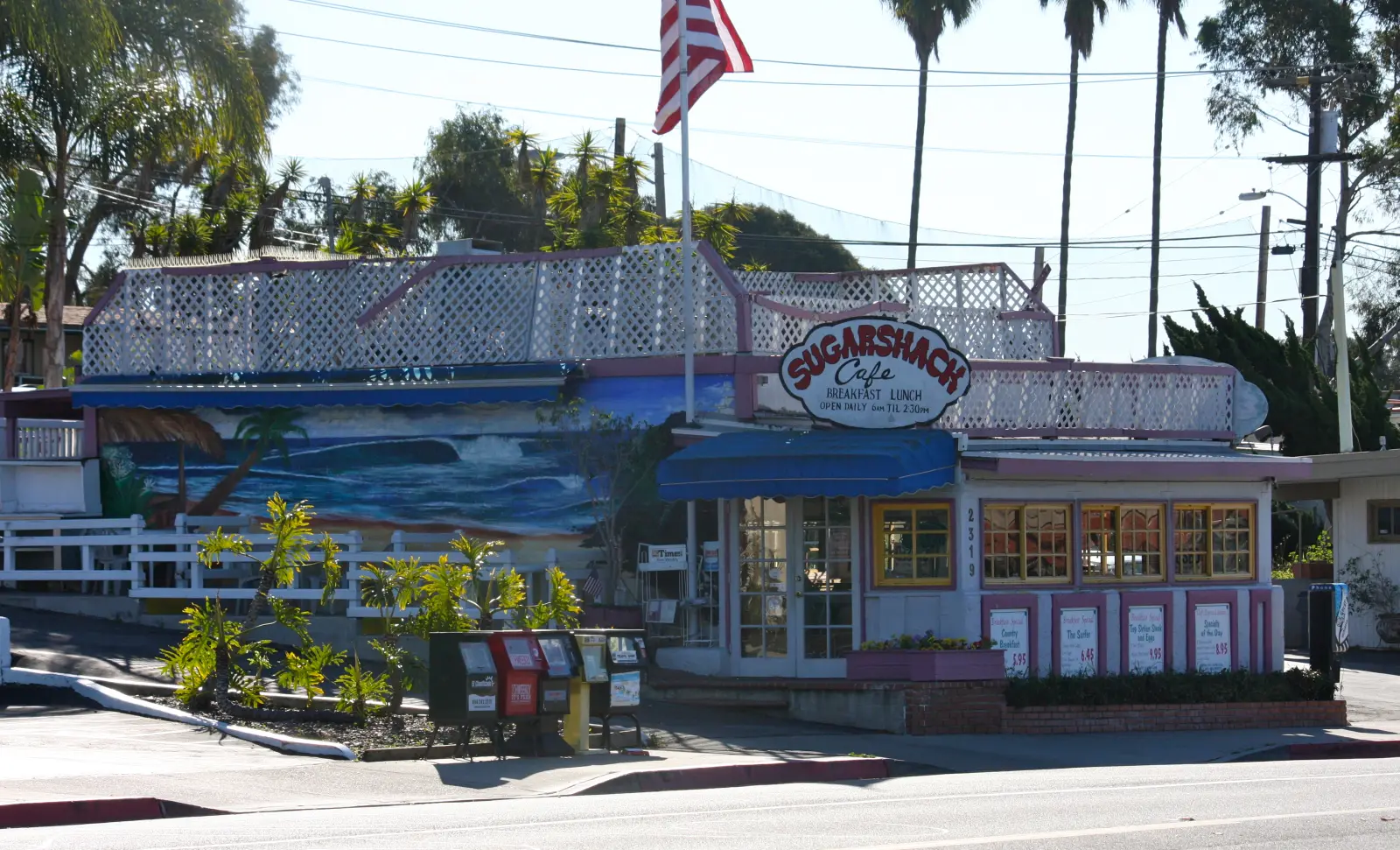 Wide sunny exterior of Rose's Sugar Shack Cafe showing the ocean-wave mural, American flag and palm trees