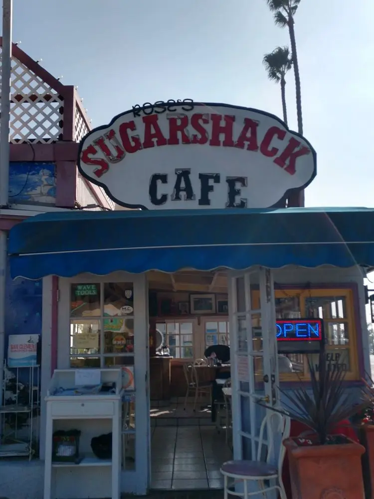 Rose's Sugarshack Cafe hand-painted sign above the blue awning with a glowing OPEN sign and palm trees behind
