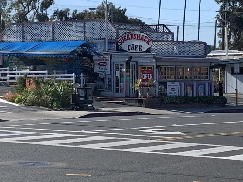 Wide sunny street view of Rose's Sugarshack Cafe with its oval sign, OPEN banner and blue awning across the crosswalk
