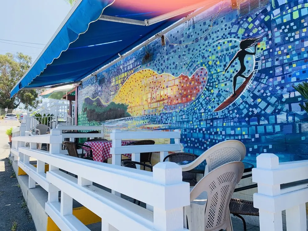 Outdoor patio under a blue awning beside a colorful mosaic mural of a surfer riding a wave, with gingham-topped tables