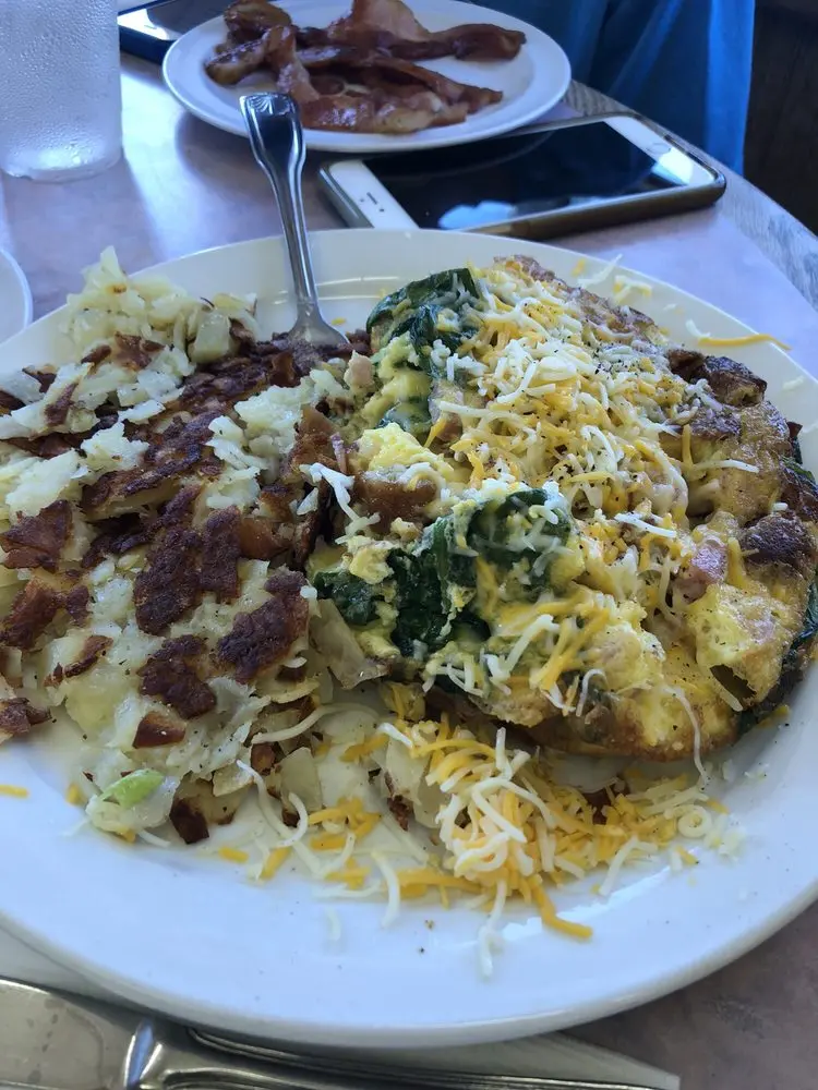 Spinach and cheese omelet topped with shredded cheese, served with hash browns and a plate of bacon behind