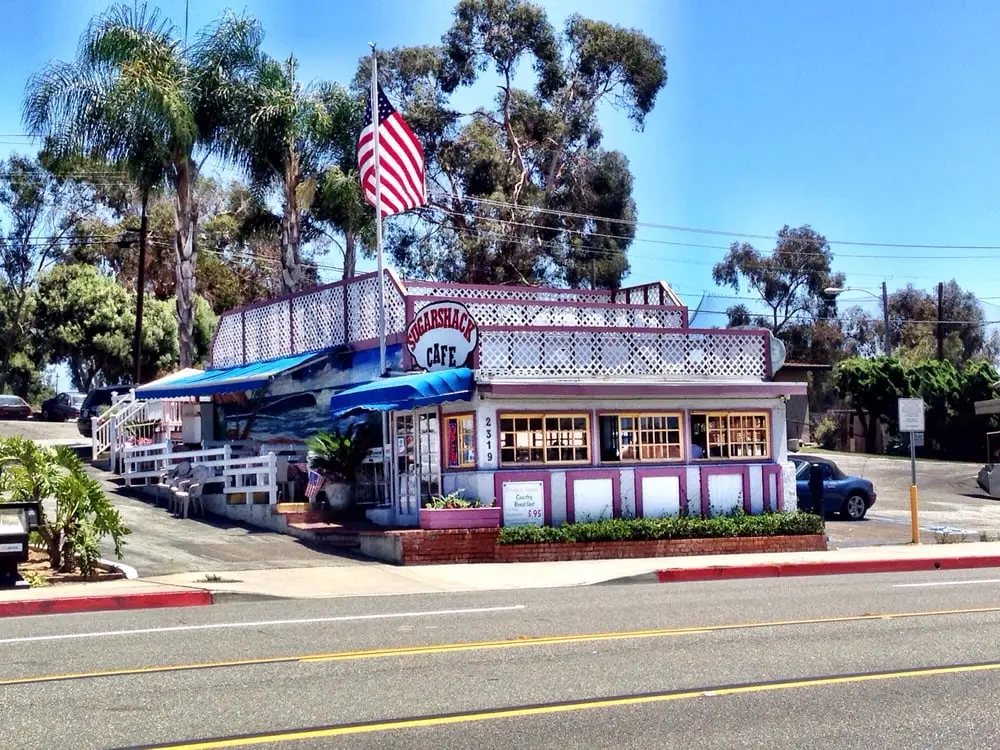 Sunny exterior of the Sugar Shack Cafe with its rooftop sign, American flag and palm trees against a bright blue sky