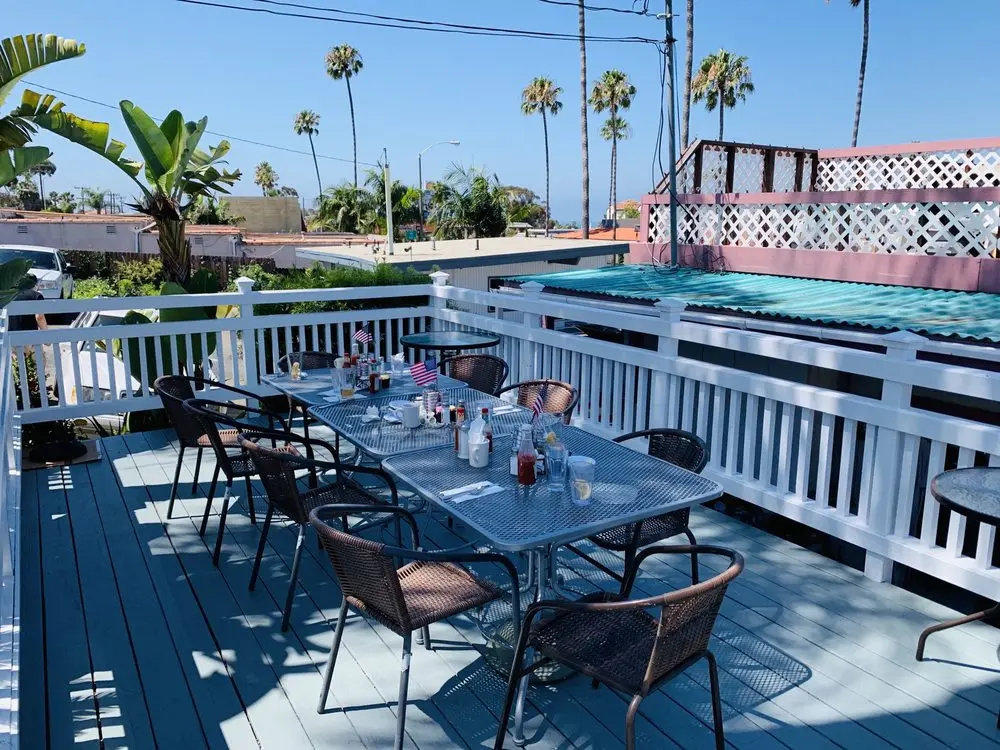 Sunlit rooftop patio deck at the cafe set with tables and chairs, framed by palm trees and a coastal town view