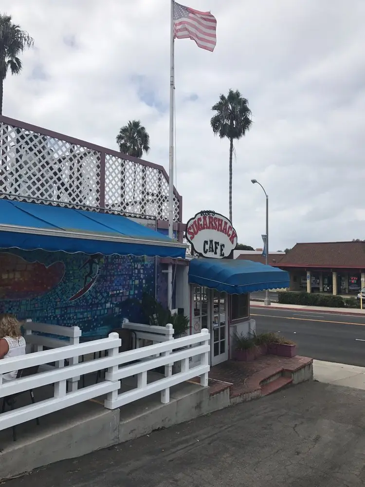 Front entrance of the Sugar Shack Cafe with its sign, blue awning and mosaic wave mural beneath palm trees