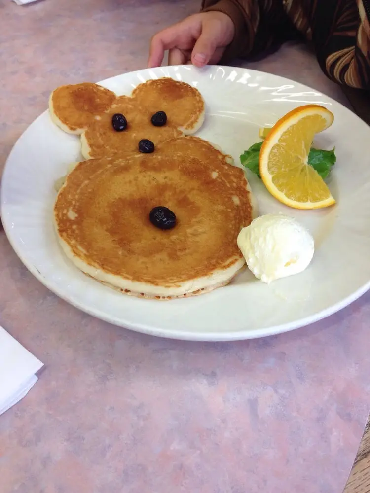Teddy-bear-shaped kids pancake with blueberry eyes and nose, topped with butter and served with an orange slice
