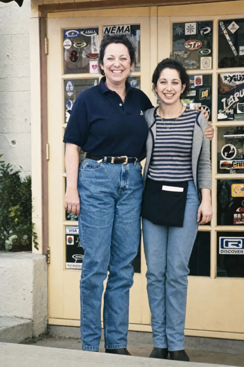 Rose and Christina, the mother-and-daughter owners of Rose's Sugar Shack Cafe, standing arm in arm in front of the cafe's sticker-covered door — restored photograph from the cafe's early years