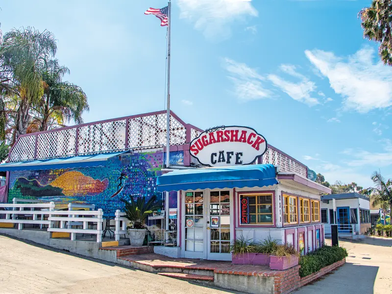 Rose's Sugar Shack Cafe storefront with its surfboard sign, beach mural and American flag under a clear blue sky